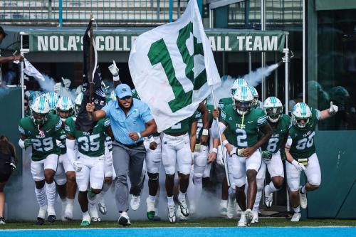 Green-uniformed football team charges from a tunnel, led by a coach, holding a flag with "12."